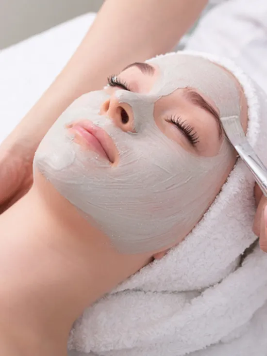 Woman receiving facial treatment with gray clay mask applied by beautician